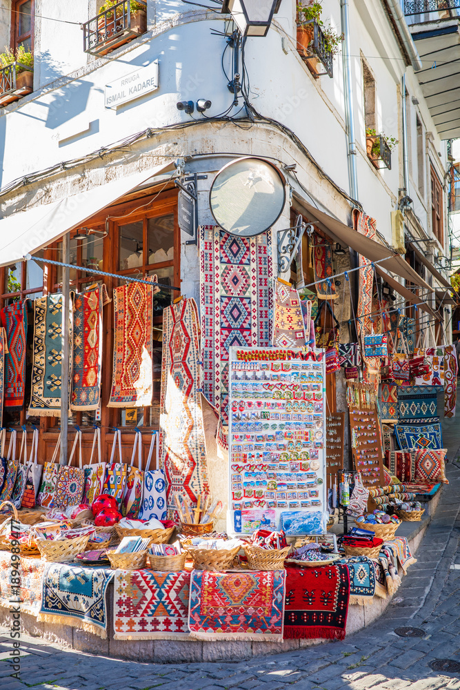 Fototapeta premium Vibrant corner shop in a European old town displaying handmade woven rugs, patterned textiles, magnets, bracelets, and cultural souvenirs outside a charming white stone building with balconies