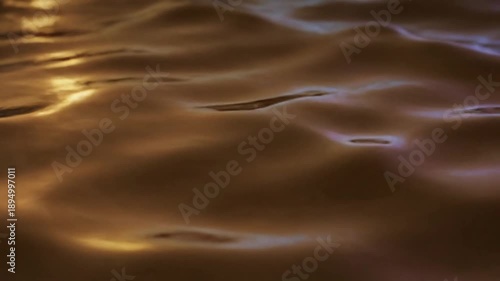 Close-up abstract view of warm golden liquid surface with light reflecting and creating flowing patterns, conveying richness and fluidity.