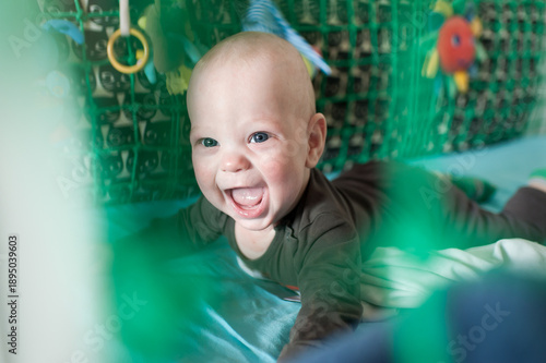 A baby is crawling on the floor and smiling widely. The background shows toys and a colorful play area. The light is bright and suggests it is daytime.