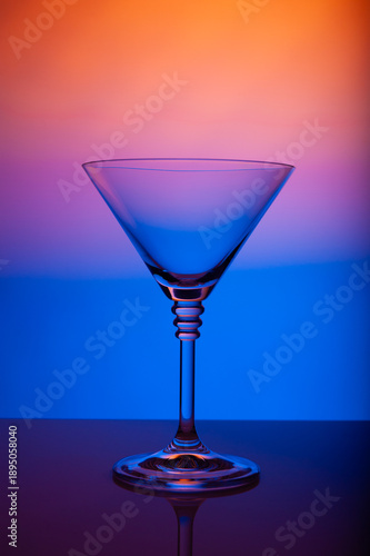 Elegant empty martini glass illuminated by colorful gradient background, reflection on glossy surface, premium cocktail glassware, modern bar still life, stylish nightlife concept