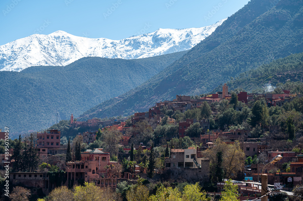 Obraz premium Mountains of Atlas loom over village in Morocco with rich greenery and snow-capped peaks