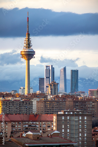 Wide suburban view of Madrid’s residential district, satellite‑covered rooftops and skyline in the background beneath grey clouds.
