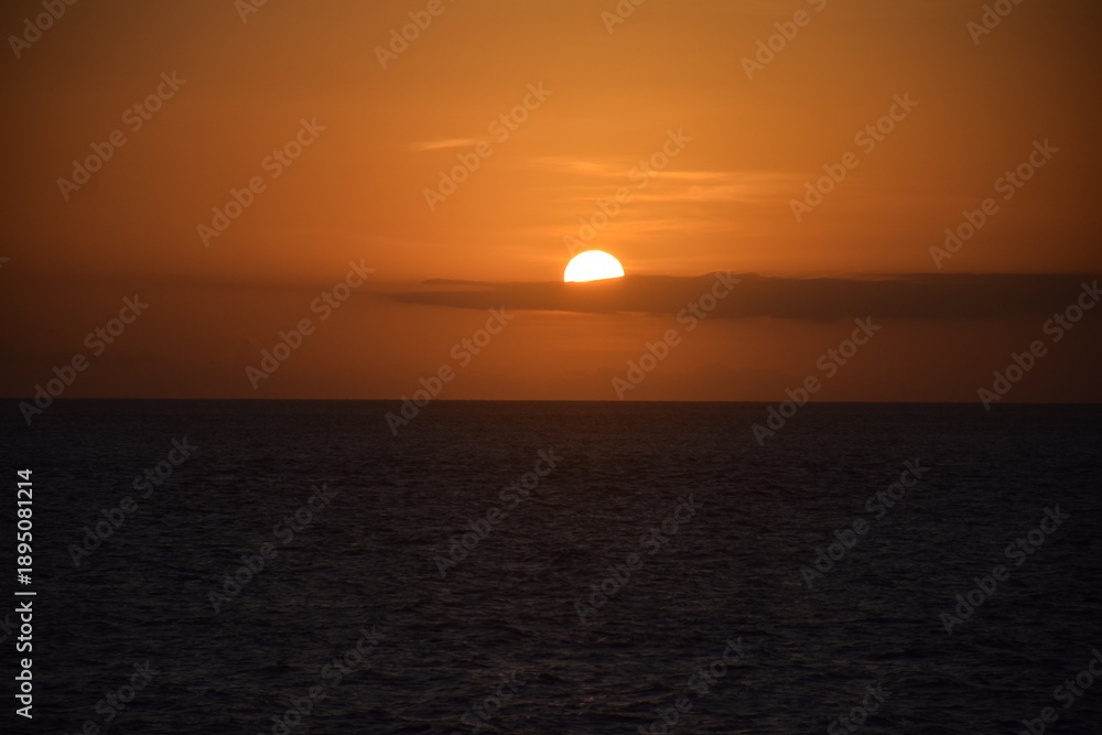 Fototapeta premium The sun appears round, surrounded by long, black clouds, and the orange sky is setting as the sun sets on the western horizon.