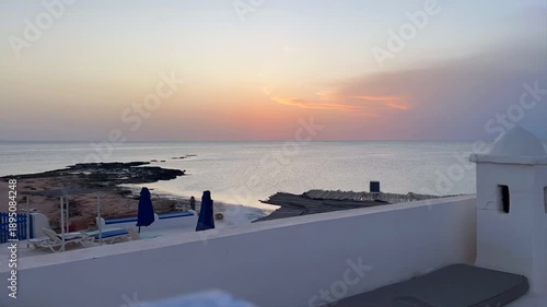 Enjoy the stunning scenery from a cozy rooftop in Djerba, Tunisia. The whitewashed terrace features blue seating, offering a perfect view of the sea and nearby palm trees.