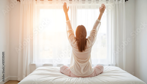 Woman stretching with arms raised on bed in bright bedroom morning  