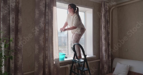 Housewife standing barefoot on windowsill, using a rag and a bucket of water to wash window glass in the room. Routine spring cleaning and household chores for maintaining cleanliness.