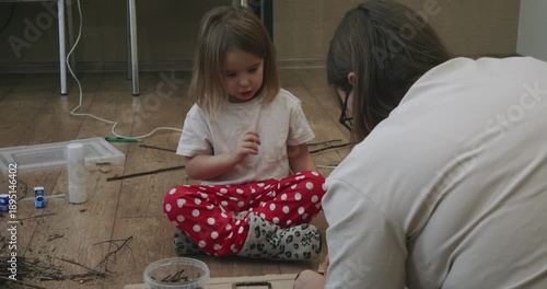 Caring mother helping her young daughter make a craft project from twigs and sticks while sitting together on the floor, fostering creativity and developing fine motor skills through play.