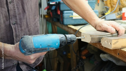 Close-up of carpenter's hands with blue screwdriver and wooden curved cut piece or furniture fragment on work surface while drilling holes on end face of piece in workshop
