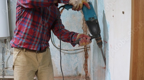 A man in brown pants and a red checkered shirt holds a drill and with great effort chisels out a long vertical groove in the wall for hiding wires, chiselling a groove in the structure of the wall