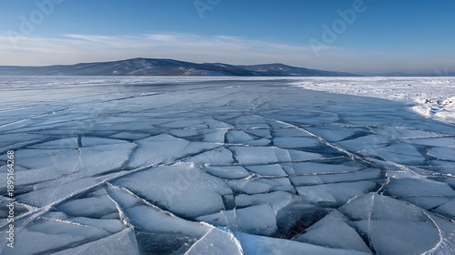 Wide angle view of a frozen lake with large ice fractures under a clear blue sky and distant hills.