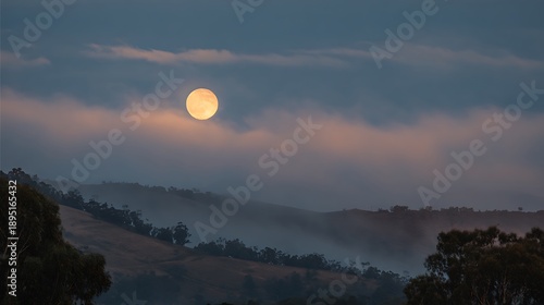 Bright full moon peeking through soft, glowing clouds over rolling hills and scattered trees.