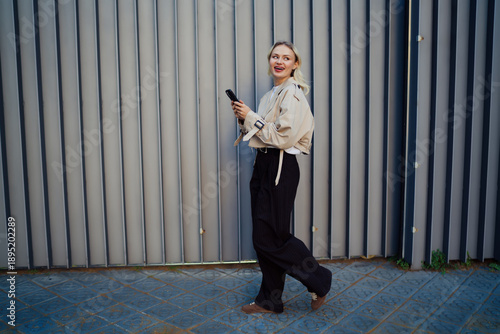 Smiling young woman walking with smartphone in hand, glancing sideways mid-step. Dynamic moment of modern connectivity and real-time interaction in motion