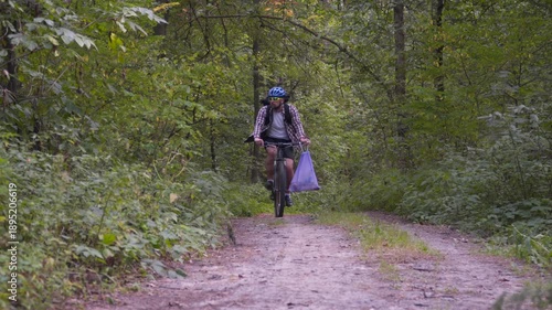 Cyclist stops on a forest trail, shows a plastic bag full of collected trash, and continues biking. Concept of plogging, environmental cleanup, nature care, and sustainable sport
