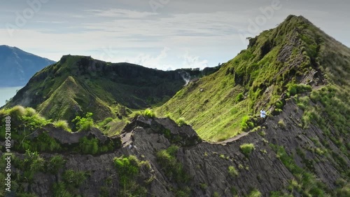 Mount Batur, Bali: Man active hiking narrow green mountain ridge connecting volcanic peaks, enjoy the challenge adventure with crater steaming in background and a vast landscape. Aerial drone flight