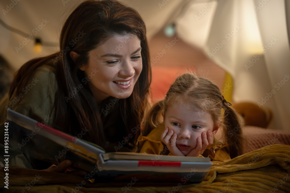 Obraz premium Mother and young daughter reading a bedtime story together under a cozy blanket fort, sharing warmth, curiosity, and affection in a calm, intimate family moment at home
