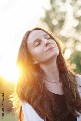 Portrait calm, beautiful young woman with closed eyes smiling at evening sunset, relieving stress after hard day in park. Serene brunette woman relaxes in the fresh air, enjoying the peace and quiet.