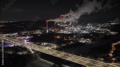 Zoomed-in nighttime drone shot of an industrial facility in Vilnius, Lithuania, with tall chimneys emitting large plumes of steam, illuminated by red warning lights over a snowy landscape.