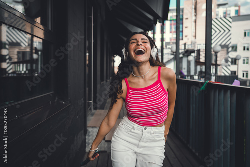 Fotografia Carefree woman laughs in headphones with raised arms, enjoying music and movement in a bright outdoor scene