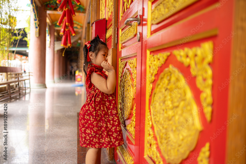 Obraz premium Adorable Little Girl in Traditional Red Cheongsam Standing by Golden Carved Temple Doors,A cute young Asian girl wearing a festive red Chinese dress (Qipao) with gold floral patterns,