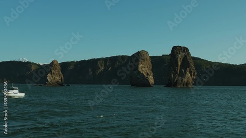 View of Three Brothers rocks located at entrance to Avacha Bay. Tourists admire landmarks from yacht and coastline with mountains and volcanoes. Slow motion