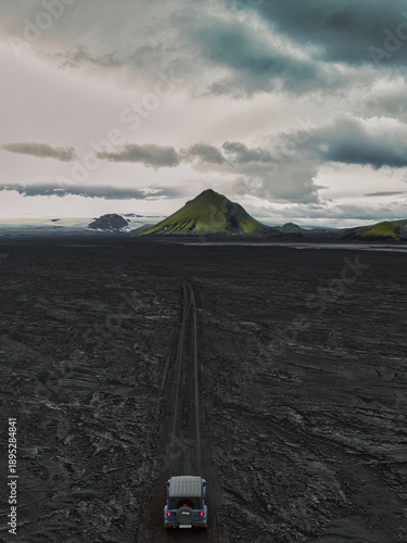Aerial view of a lone car threads a stark path through the volcanic expanse towards the verdant, pyramidal peak of Maelifell, Skaftarhreppur, Iceland.