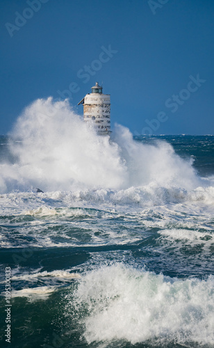 Powerful storm waves crashing against an isolated lighthouse standing in rough open sea conditions.