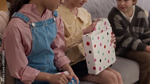 Close up of two birthday biracial girls sitting on sofa and opening presents while their friends watching them