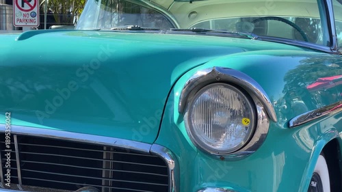 A turquoise classic car is shown in close up with smooth bodywork and chrome trim gleaming in the sun. The vintage headlight and windshield reflect trees and nearby show tents.
