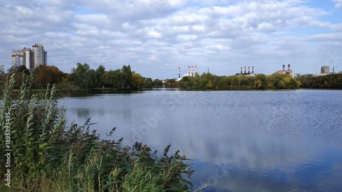 Trees with yellow leaves grow on the grassy banks of the river. On the opposite shore there are living houses, administrative buildings, and factory chimneys. The reeds are in the water. Sunny autumn