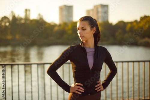 Woman runner resting by river after training. Hands on hips, the female athlete pauses on a riverside path at dusk, cooling down and showing confidence. Recovery and focus sport fitness concept.