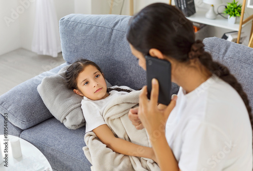 Sick tired child lying on sofa at home, mother calling to doctor for advice on treatment for colds and flu. Parent speaking by phone with pediatrician about fever while giving care to daughter