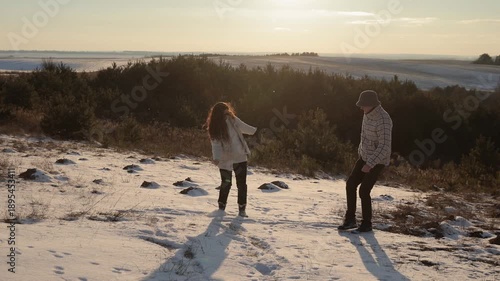 Active young family plays kicking snow and hugs on hill slope at sunset highland. Excited man and woman have fun together in winter weather. Romantic date in nature at cold season