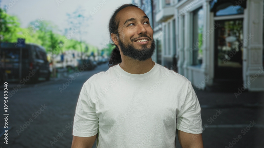 Fototapeta premium Man in white t-shirt smiling and looking up on a city street with storefronts and parked cars visible; optimism urban calm.