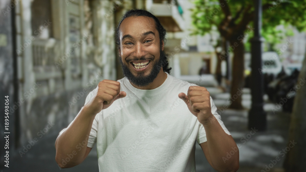 Fototapeta premium Bearded man with hands raised and clenched fists on a city street smiling widely and looking ahead in urban outdoor setting wearing a white t shirt; joy celebration energy.