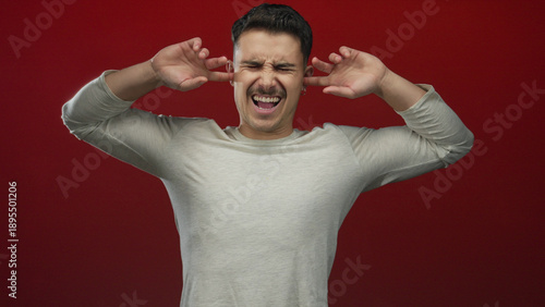Cuadro en lienzo Young hispanic man covers ears against a noisy red backdrop, expressing annoyance and isolation