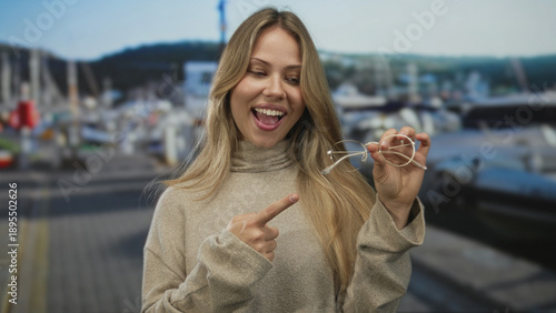 Photography Blonde woman smiling while holding eyeglasses and pointing finger to object at seaside port; demonstration highlight