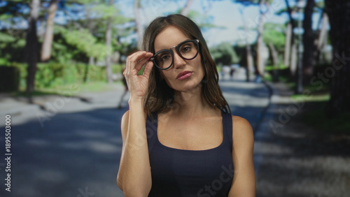 Foto Young hispanic woman adjusts round black glasses on a tree-lined street at midday; quiet contemplation