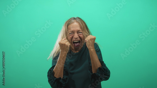 Foto Senior woman blonde with clenched fists and mouth open, head tilted back, wearing teal blouse in studio; excitement celebration