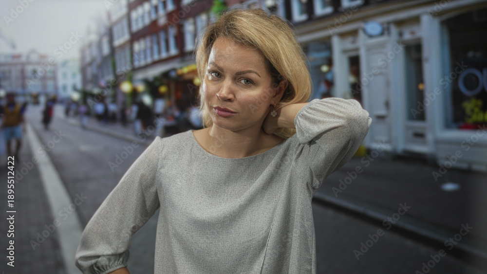 Fototapeta premium Young blonde woman lightly touches her neck outside a brown brick building on a busy street; contemplation.