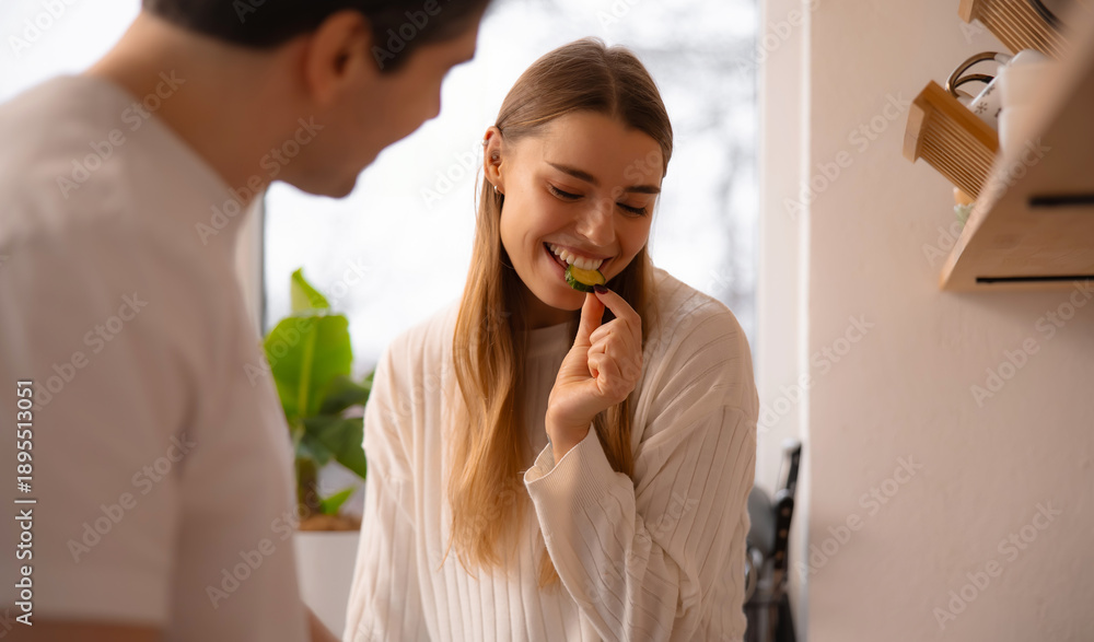 Fototapeta premium Happy woman enjoying a cucumber slice while her partner cooks in the background, bright kitchen lifestyle and healthy eating theme.
