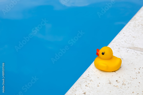 Close-up of a yellow rubber duck sitting on the right pool edge, looking into blue water with white cloud reflections. Concept of summer vacation, freelance. Copy space on the left