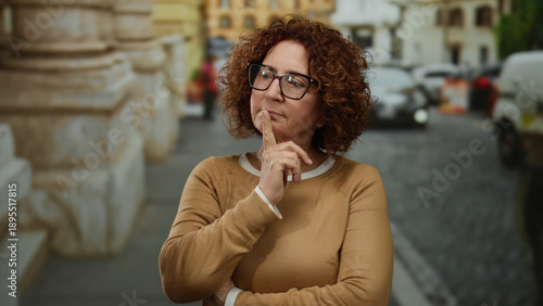Foto Woman pondering on a city street, wearing glasses and brown sweater, with cars and buildings in the blurred background during daytime