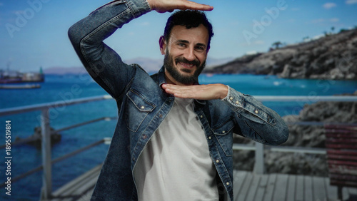 Hispanic man with beard, in denim shirt, making a gesture by the beach with the sea and mountains in the background, capturing a relaxed and joyful outdoor moment.