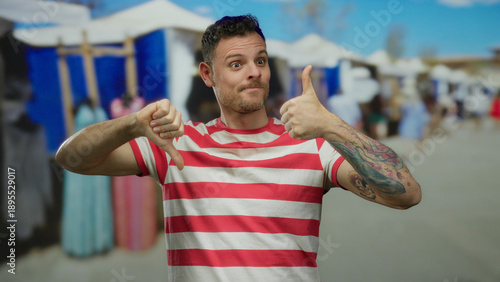 Foto Man wearing striped shirt outdoors gives thumbs up and down expressing mixed feelings in urban marketplace setting