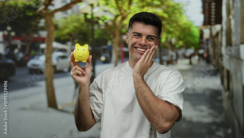 Young man smiling holding yellow piggy bank on busy city street with greenery around, capturing a joyful outdoor moment showcasing savings and urban lifestyle.