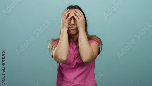 Fotografie Woman distressed, hands on face over blue background, expressing frustration and worry, wearing a pink shirt, isolated and emotional, conveying stress and concern