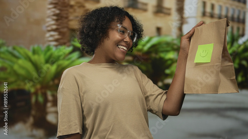 Εκτύπωση καμβά Woman wearing glasses holds paper bag with smiley note on city street; happiness generosity kindness