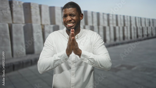 Fotografie Man in white shirt with pressed palms praying gesture on street by stacked concrete blocks; hopeful prayer