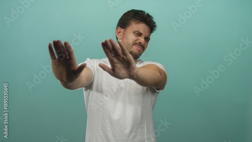 Young hispanic man palms out stop gesture, bare hands and face visible, studio teal backdrop; repulsion refusal avoidance.
