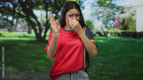 Woman holds nose and grimaces wearing a spanish soccer jersey in a park, pinching nose toward camera; disgust reaction.